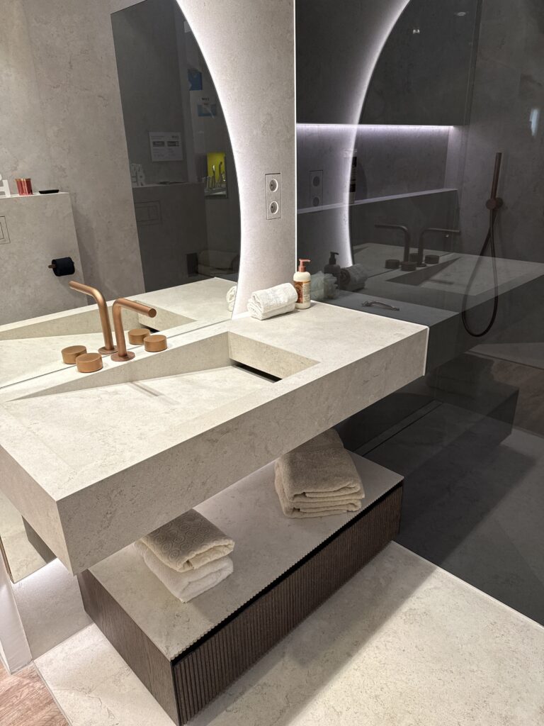 Ausstellungsbad mit Naturstein-Waschtisch, kupferner Wandarmatur und rundem beleuchtetem Spiegel